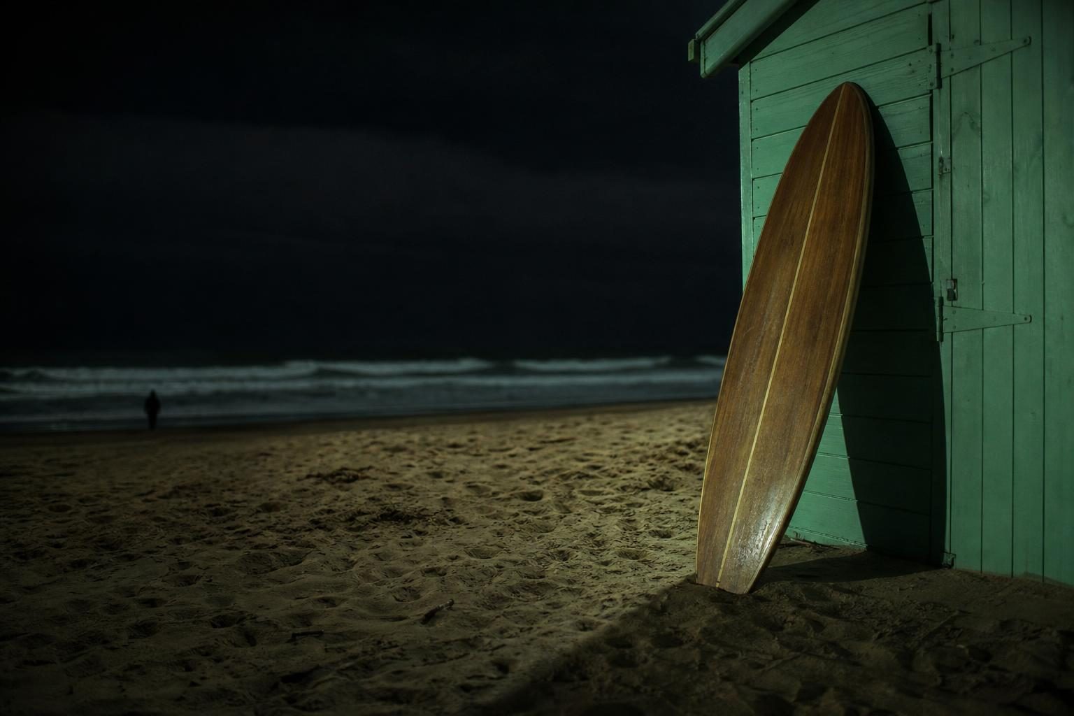 A lone wooden surfboard leans against a brightly painted beach hut, casting a long shadow on the golden sand as the ocean waves crash rhythmically in the background.
